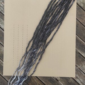 May include: A bundle of 10 black and white synthetic dreadlocks, 50 inches long. The dreadlocks are displayed on a brown paper background with a label that reads "REF: NB 04 Lot de 10 Longueur: 50 cm".