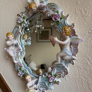 Puede incluir: Espejo ovalado con marco azul claro y blanco, adornado con figuras de querubines y rosas rosas. El marco tiene detalles dorados. El espejo refleja una lámpara de araña y otros objetos. Una pieza decorativa para un dormitorio o sala de estar.