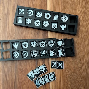 Könnte beinhalten: Zwei schwarze Tabletts mit weißen Symbolen, wahrscheinlich für ein Brettspiel, liegen auf einer Holzoberfläche. Die Tabletts enthalten verschiedene Icon-Kacheln. Mehrere lose Kacheln mit passenden Symbolen sind ebenfalls vorhanden.