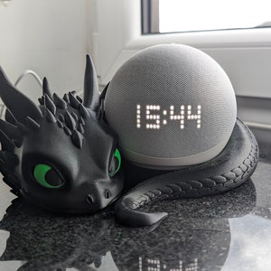 Könnte beinhalten: Ein schwarzer Lautsprecherhalter in Drachenform mit grünen Augen hält einen grauen Smart-Lautsprecher, der die Uhrzeit 15:44 anzeigt. Der Halter ist als Drache gestaltet und verleiht dem Lautsprecher eine dekorative Note.