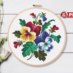 Puede incluir: Un patrón de punto de cruz con un ramo de pensamientos coloridos en tonos de amarillo, rojo, azul y verde. El patrón se muestra en un aro de bordado de madera.