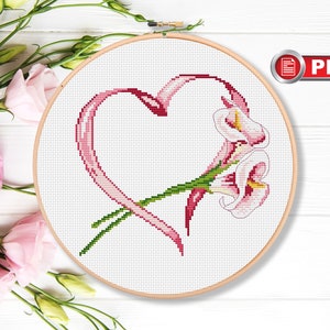 Pode incluir: Padrão de ponto cruz com uma fita rosa em forma de coração com uma flor branca e rosa dentro. O padrão está em um aro de bordado de madeira.