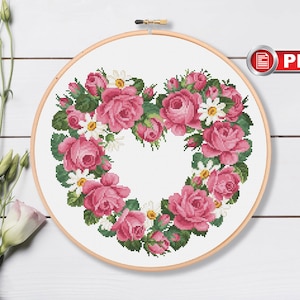 Könnte beinhalten: Ein Kreuzstichmuster einer herzförmigen Girlande aus rosa Rosen und weißen Gänseblümchen. Das Muster ist in einem Reifen und bereit zum Sticken.