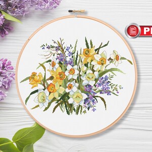 May include: A cross stitch pattern featuring a bouquet of white and yellow daffodils with purple wildflowers. The pattern is displayed on a white background in a round embroidery hoop.