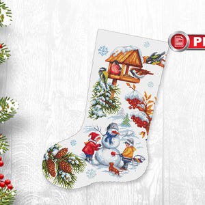 Puede incluir: Una media de Navidad blanca con una escena de invierno que presenta un muñeco de nieve, dos niños, pájaros y un comedero para pájaros. La media está decorada con piñas y copos de nieve.