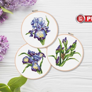 May include: Three circular wooden embroidery hoops containing cross-stitch patterns of purple and blue iris flowers with green stems and leaves on white fabric. A red and white PDF icon is visible in the upper right corner of the image.