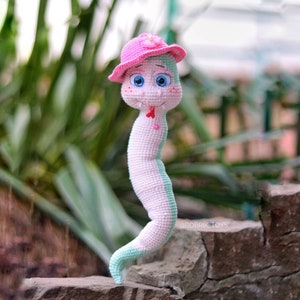 May include: A crocheted snake toy wearing a pink hat with a flower. The snake is pink and green and has a friendly face with big blue eyes.