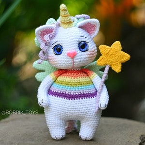 Op de afbeelding: Een witte gehaakte eenhoornkat met een regenboog gestreept shirt, groene vleugels en een gouden sterrewand. De kat heeft blauwe ogen en een roze neus.