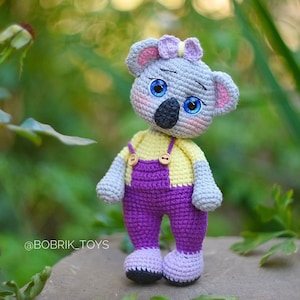 Puede incluir: Un juguete de koala de crochet que lleva un mono morado con detalles amarillos y un lazo rosa. El juguete tiene ojos azules y una cara gris.