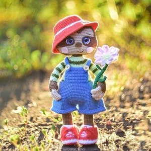 Puede incluir: Un perezoso de crochet que lleva un sombrero rojo, un mono azul y una camisa a rayas, sosteniendo una flor morada. El perezoso está de pie sobre una superficie marrón con plantas verdes en el fondo.