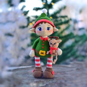 Peut inclure: Une poupée elfe en crochet vert et rouge tenant un petit renne en crochet. L'elfe porte un chapeau blanc avec un pompon, une chemise à rayures vertes et rouges et des bottes brunes. Le renne porte une écharpe rouge.