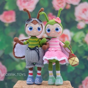 May include: Two crocheted toy bugs, one green and one brown, with large eyes and smiling faces. The green bug is wearing a pink dress and holding a small lantern. The brown bug is wearing a green and yellow striped shirt and gray pants. The bugs are standing on a wooden surface with a blurred background of pink roses.