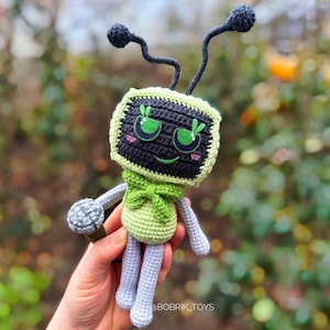 Op de afbeelding: Een groen en grijs gehaakt robot speelgoed met een microfoon. De robot heeft een vierkant hoofd met een zwart scherm en groene ogen. De robot draagt een groene strik.
