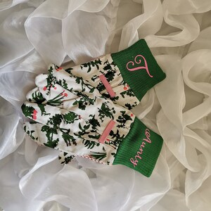 Peut inclure: Une paire de gants de jardinage blancs avec des poignets verts et des lettres brod&eacute;es roses. Les gants ont un motif floral avec des feuilles vertes et des fleurs blanches.