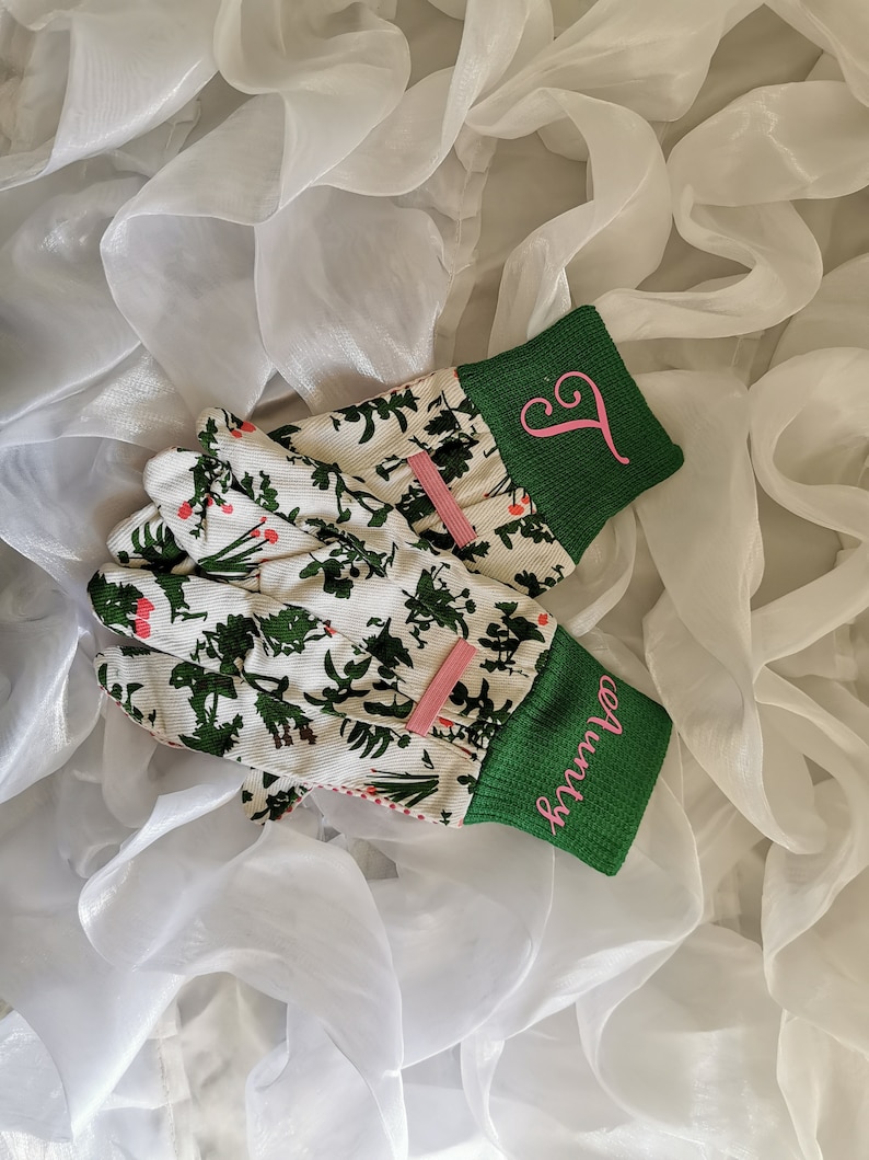 Peut inclure: Une paire de gants de jardinage blancs avec des poignets verts et des lettres brod&eacute;es roses. Les gants ont un motif floral avec des feuilles vertes et des fleurs blanches.