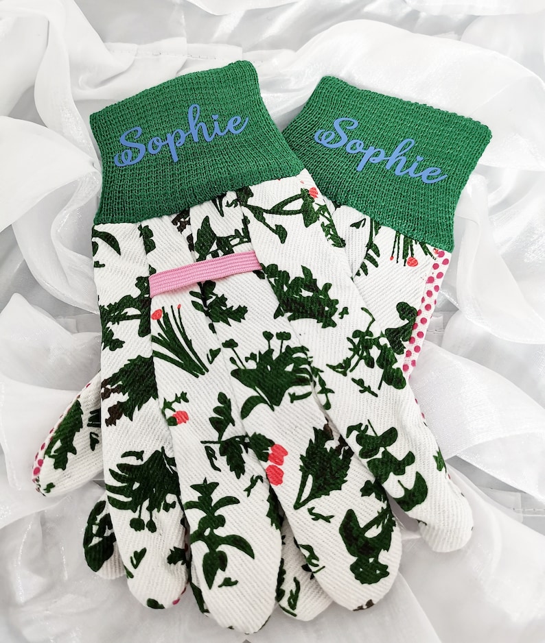 Peut inclure: Une paire de gants de jardinage blancs avec un poignet vert et un motif floral. Les poignets sont personnalis&eacute;s avec le nom "Sophie" en lettres bleues.