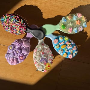 May include: Five decorative hair brushes with various designs. One brush is covered in rainbow sprinkles, others feature cute embellishments. The brushes have different coloured handles. Each brush is approximately 15 cm long.