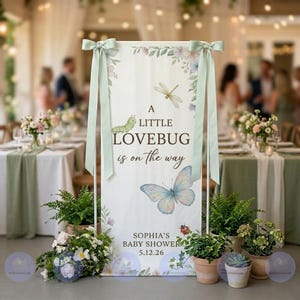 May include: A white rectangular sign with the text "A LITTLE LOVEBUG is on the way" and "SOPHIA'S BABY SHOWER 5.12.26". The sign features illustrations of a caterpillar, dragonfly, butterfly, and ladybug. The sign is decorated with floral accents and tied with light green ribbons.