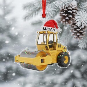 May include: A yellow road roller Christmas ornament with a Santa hat that says "LUCAS". The ornament is decorated with a string of colorful lights and has a red ribbon for hanging. The background is a snowy scene.