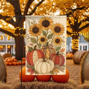 May include: A rectangular garden flag featuring a printed illustration of three pumpkins and a bouquet of sunflowers. The pumpkins are orange, red, and white with various patterns. The sunflowers have yellow petals and brown centers.