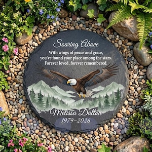 May include: A round slate memorial stone featuring a bald eagle in flight over a mountain and forest landscape. The text reads: Soaring Above, With wings of peace and grace, you’ve found your place among the stars. Forever loved, forever remembered. Melissa Dalton 1979-2026.