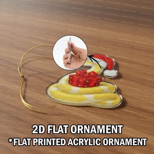 May include: A flat, acrylic Christmas ornament depicting a yellow and white snake wearing a Santa hat and holding a red gift box. The ornament has a gold string for hanging. Text at the bottom reads "2D FLAT ORNAMENT" and "*FLAT PRINTED ACRYLIC ORNAMENT."