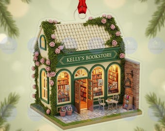 May include: A miniature bookstore ornament with the text "KELLY'S BOOKSTORE". The building features a green and white facade, a tiled roof, and is adorned with pink roses and greenery. The interior is lit, showcasing bookshelves and furniture.