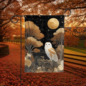 May include: A garden flag featuring a nighttime scene with a full moon and a white and brown owl perched on a branch. The flag has a black background with gold-colored trees and foliage. The flag is displayed on a black metal stand in an outdoor setting.
