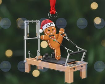 Könnte beinhalten: Ein Lebkuchenmann-Weihnachtsschmuck mit einem Weihnachtsmannhut mit dem Namen "Riley", der auf einem Pilates-Reformer-Gerät trainiert. Der Schmuck ist braun, rot und weiß, mit einem roten Band zum Aufhängen.