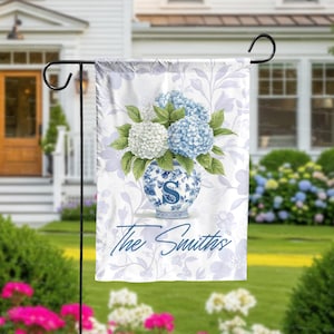 May include: A garden flag featuring a blue and white floral design with a vase of hydrangeas. The vase has the letter "S" on it. The words "The Smiths" are written in script below the vase.