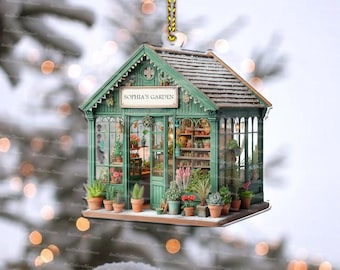 May include: A green and white Christmas ornament shaped like a miniature greenhouse with the words "Sophia's Garden" on the front. The ornament is decorated with faux plants and flowers.