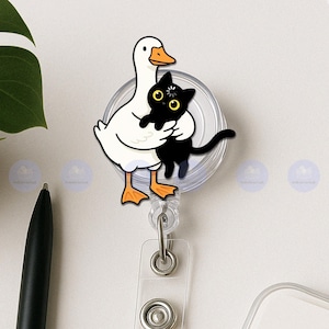 Puede incluir: Un portatarjetas con un ganso de dibujos animados abrazando a un gato negro. El ganso es blanco con patas y pico naranjas. El gato tiene ojos amarillos y una pequeña flor en la cabeza. El carrete es transparente con un cordón retráctil y un clip metálico.