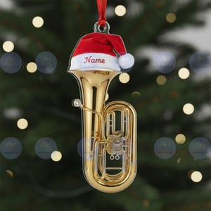 May include: A gold-colored tuba Christmas ornament with a red Santa hat. The hat has a white brim and a white pom-pom. The word "Name" is printed on the hat. A red ribbon is attached for hanging.