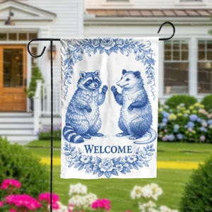May include: A white garden flag featuring a blue illustration of a raccoon and an opossum waving. The flag has a floral border and the word "WELCOME" in blue. The flag is displayed on a black metal stand in a garden setting.