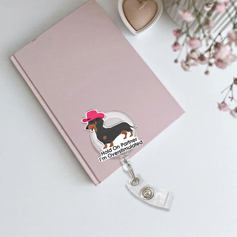 May include: A clear badge reel with a cartoon dachshund wearing a pink cowboy hat. The dachshund is black and brown. The text on the badge reel says "Hold On Partner I'm Overstimulated". The badge reel is attached to a pink notebook.