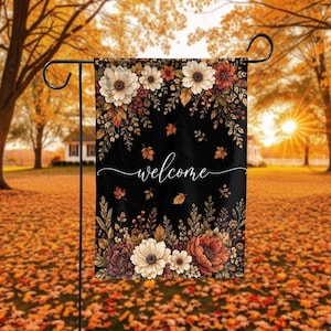 May include: A rectangular garden flag featuring a black background with a floral border of white, orange, and deep red flowers and autumn leaves. The word welcome is printed in white cursive script in the center of the flag, which hangs on a black metal stand.