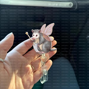 May include: A whimsical opossum badge reel featuring a fairy opossum with purple wings and a star wand. The opossum is gray and white, with a cute expression. The badge reel has a clear plastic clip and a retractable cord.