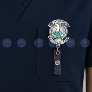 May include: A navy blue scrub top with a pocket, featuring a badge reel. The badge reel has a cartoon goose wearing a medical coat and holding a scalpel. Text on the badge reads "Here's the thingy with the pointy end."