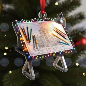 Könnte beinhalten: Ein Weihnachtsornament in Form eines Architektentisches. Der Tisch zeigt einen Bauplan mit dem Wort "NAME". Buntstifte und eine kleine Lampe befinden sich auf dem Tisch. Der Tisch ist mit bunten Lichtern verziert und hat ein rotes Band zum Aufhängen.