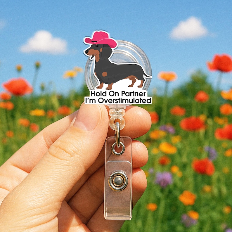 May include: A retractable badge reel featuring a cartoon dachshund wearing a pink cowboy hat. The text "Hold On Partner I'm Overstimulated" is printed on the badge. The reel has a clear plastic clip and a metal snap.