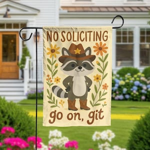 May include: A garden flag with a raccoon wearing a cowboy hat and boots, surrounded by flowers. The flag reads "NO SOLICITING go on, git" in brown text on a cream background. The flag is hanging on a black metal stand.