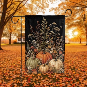May include: A rectangular garden flag featuring a black background with a central arrangement of four pumpkins in shades of orange, green, and cream, surrounded by a variety of wildflowers, dried grasses, and autumn foliage in warm tones.