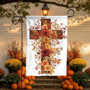 May include: A white garden flag featuring a cross design composed of autumn-colored flowers and foliage. The cross is filled with red, orange, and yellow blooms, with detailed leaves and branches. The flag is displayed on a black metal stand.
