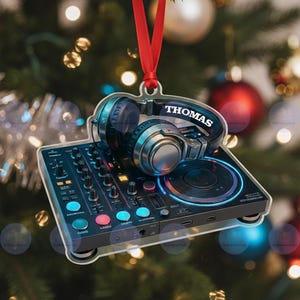 Puede incluir: Adorno navideño de acrílico transparente con una mesa de mezclas de DJ y auriculares. Los auriculares son plateados y negros, con el nombre "THOMAS" impreso. La mesa de mezclas tiene botones y luces de colores, y una cinta roja para colgar.