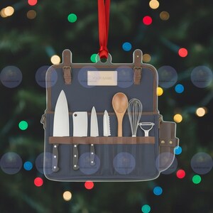 May include: A clear acrylic ornament shaped like a chef's knife roll. The navy blue roll features a variety of kitchen tools, including knives, a wooden spoon, and a whisk. The text "YOUR NAME" is printed on a small label. A red ribbon is attached for hanging.