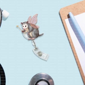May include: A badge reel featuring a whimsical opossum fairy with pink and brown wings, holding a golden wand. The opossum is gray and white. The reel has a clear plastic clip and a retractable cord.