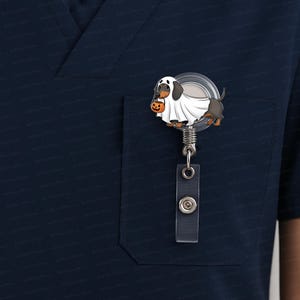 Puede incluir: Un carrete retr&aacute;ctil para placa con tem&aacute;tica de Halloween con un perro salchicha disfrazado de fantasma, sosteniendo una calabaza. El carrete de la placa tiene una parte frontal de pl&aacute;stico transparente, un clip de metal y un cord&oacute;n retr&aacute;ctil. El perro es negro, marr&oacute;n y blanco.