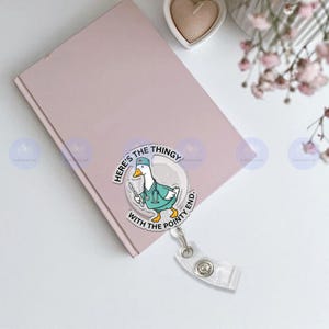 May include: A pink notebook with a badge reel featuring a cartoon duck wearing a medical uniform and holding surgical tools. The badge reel has the text "Here's the thingy with the pointy end."