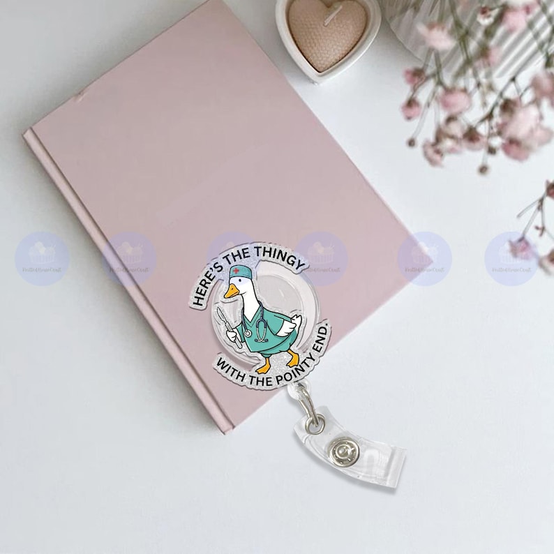 May include: A pink notebook with a badge reel featuring a cartoon duck wearing a medical uniform and holding surgical tools. The badge reel has the text "Here's the thingy with the pointy end."