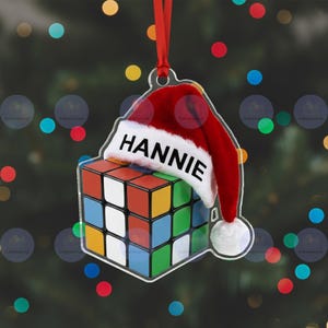 May include: A clear acrylic Christmas ornament featuring a Rubik's Cube design with a red Santa hat. The hat has the name "HANNIE" printed on it. The cube has red, blue, green, orange, yellow, and white squares. A red ribbon is attached for hanging.